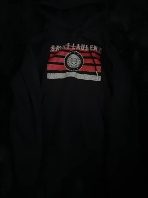 Svart hoodie från Saint Laurent - Svart hoodie från Saint Laurent med tryck i rött, vitt och svart framtill. Klassisk huva med dragsko och en cool logga på bröstet. Perfekt för dig som vill ha en streetstyle-look med lyxig känsla.