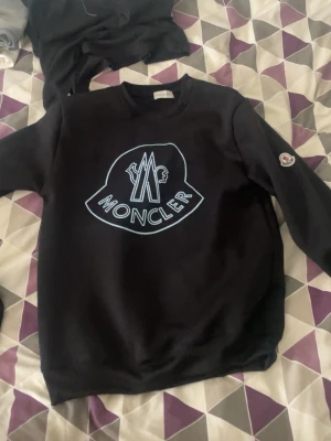 Svart Moncler hoodie med logga - Svart hoodie från Moncler med stor vit logga framtill och Moncler-märke på ena ärmen. Klassisk passform med rund hals och långa ärmar. Perfekt för dig som gillar streetwear och vill sticka ut med en snygg designertröja.