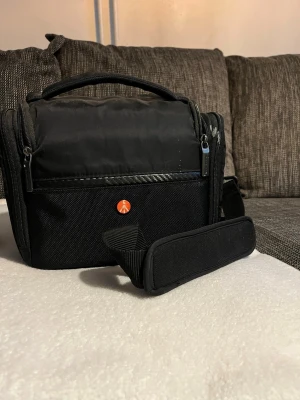 Svart datorväska från Manfrotto - Snygg svart kameraväska från Manfrotto med praktiskt handtag och justerbar axelrem. Väskan har flera fack med dragkedja och detaljer i kolfiber-look. Perfekt för att bära kamera och tillbehör. Tillverkad i slitstarkt textilmaterial.