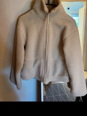 Beige teddyjacka med dragkedja - Mysig beige teddyjacka med hög krage och hel dragkedja framtill. Jackan har en avslappnad passform och är tillverkad i fluffigt teddy-material som håller dig varm. Perfekt för kyliga dagar och enkel att matcha med olika outfits.