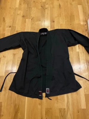 Svart kampsportsjacka Budo-Nord stl 160 cm - Säljer en svart kampsportsjacka från Budo-Nord, perfekt för karate, judo eller annan kampsport. Jackan är i kraftigt bomullstyg med förstärkta sömmar och knytband i midjan. Klassisk modell med lång ärm och öppen front.