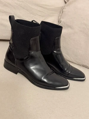 Svarta Chelsea boots med nitar - Snygga svarta Chelsea boots i blankt skinn med stickad textil upptill och coola silverdetaljer på tån och nitar längs hälen. Klassisk siluett med modern twist, perfekt för dig som vill sticka ut lite extra.