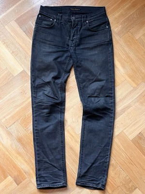 Svarta slim fit jeans från Nudie Jeans - Snygga svarta slim fit jeans från Nudie Jeans med klassisk femficksdesign och subtila slitningar. Jeansen har normal midja, raka ben och är tillverkade i mjuk denim med stretch för extra komfort. Modell Grim Tim. W:30 L:32