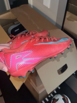 Röda Nike Mercurial fotbollsskor - Säljer ett par röda Nike Mercurial fotbollsskor med ljusblå detaljer och snörning. Skorna har en låg profil och är tillverkade i syntetmaterial för bra grepp och kontroll på planen. Perfekta för dig som vill ha snabba och lätta skor till matchen.