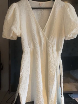 Vit omlottklänning med puffärm från Nelly  - Superfin vit kort omlottklänning med puffärmar och knyt i midjan. Klänningen har en strukturerad yta och v-ringning, vilket ger en trendig och fräsch look. Perfekt för varma dagar eller examen! Aldrig använd. Storlek 46