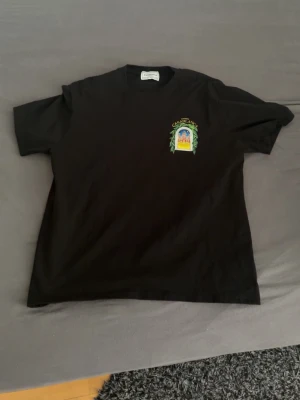 Svart t-shirt Casablanca Avenida - Svart t-shirt från Casablanca med färgglatt tryck av en byggnad och texten 'Avenida Casablanca' på ryggen. Mindre tryck på bröstet. Klassisk passform och rund hals. Perfekt för dig som gillar unika prints och streetwear vibes.