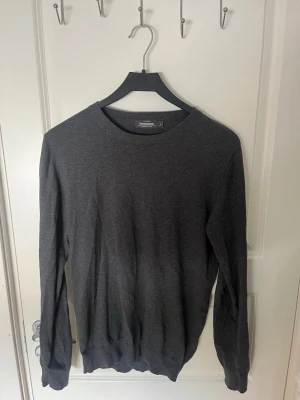 Mörkgrå tröja från Dressmann - En enkel och stilren mörkgrå tröja från Dressmann med rund halsringning och långa ärmar. Tröjan är tillverkad i ett mjukt och tunt material som passar perfekt till lager på lager och har ribbade muddar vid ärmslut och nederkant.