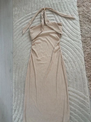 Beige långklänning med halterneck - Snygg beige långklänning med halterneck och lätt rynkad struktur. Klänningen har en smickrande passform och är ärmlös med knyt runt nacken. Perfekt för sommarens alla tillfällen.