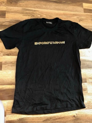 Svart Emporio Armani t-shirt - Svart t-shirt från Emporio Armani med guld logotyptryck över bröstet. Klassisk rund halsringning och korta ärmar. Perfekt för en stilren och trendig look. Skulle säga att den passar S-M. Priset kan sänkas vid snabb affär 