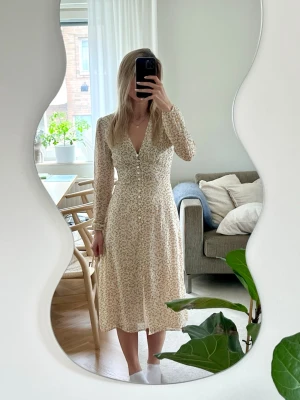 Blommig långklänning från Adoore - Romantisk långklänning från Adoore i ljusbeige med små blommor i gult och grönt. Klänningen har v-ringning, långa ärmar och knappar framtill hela vägen ner. Perfekt för sommarens alla tillfällen!                                                        Fint skick, men finns en del revor i tyget, se bilder.  Även en sträckning i tyget på ena sidan, men det är inget jag tycker man ser när man har på den, se bilder. 