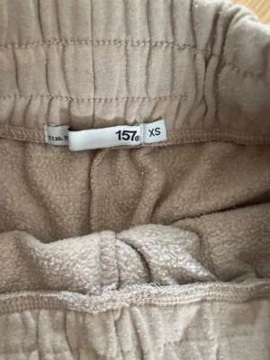 Beige mjukisbyxor från 157, XS - Beiga mjukisbyxor från 157 i storlek XS. Byxorna har raka ben, resår i midjan och två bakfickor. Mjukt och fleeceliknande material på insidan som gör dem extra sköna. Perfekta för en avslappnad stil.