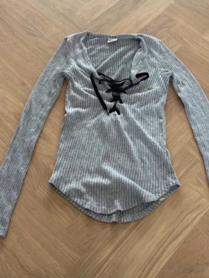 Grå ribbad topp med snörning Gina Tricot - Snygg grå långärmad topp från Gina Tricot i ribbad bomullsmix. Toppen har v-ringning med svart snörning framtill som ger en cool detalj. Perfekt till jeans eller kjol för en trendig look.
