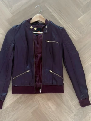 Mörklila skinnjacka med huva - Snygg mörklila skinnjacka med avtagbar huva och guldfärgade dragkedjor. Jackan har tre fickor framtill, ribbade muddar och en cool, figurnära passform. Perfekt för dig som vill sticka ut med en unik färg och detaljer.