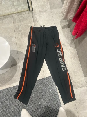 Gasp Byxor - Svarta mjukisbyxor från GASP INC med orangea ränder längs sidorna och tryck i vitt och orange. Byxorna har resår och dragsko i midjan samt tryckta detaljer på både fram- och baksida. Perfekta för chill eller gymmet.