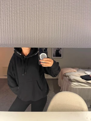 Svart hoodie med snörning i huvan - Säljer en svart hoodie med huva och snörning, samt en stor magficka framtill. Hoodien har en relaxed fit och är perfekt för en chill och avslappnad stil. Materialet känns mjukt och bekvämt, och det finns vita detaljer på snörena vid huvan.