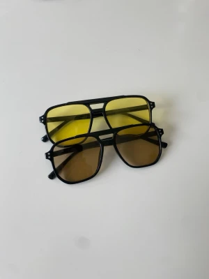 Två par retro solglasögon svart båge - Två par solglasögon med svart båge och stora rektangulära glas. Ena paret har gula linser och det andra har bruna linser. Perfekt accessoar för att lyfta din stil med en cool retrovibe.