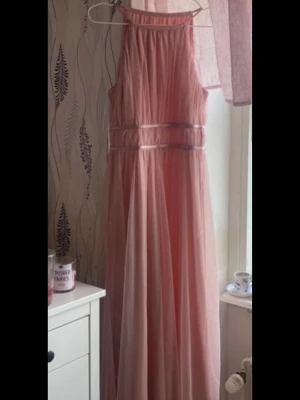 Lång rosa tyllklänning - Säljer en romantisk långklänning i ljusrosa tyll med markerad midja och ärmlös design. Klänningen har ett tunt, transparent lager över ett mjukt innerfoder och breda band i midjan som ger en elegant siluett. Perfekt för dig som vill sticka ut med en feminin look.