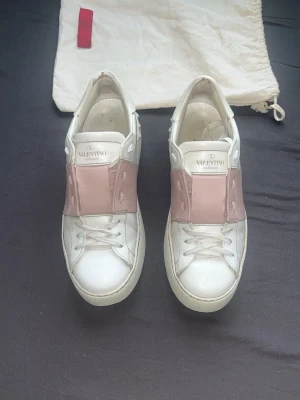 Valentino rosa - Snygga vita sneakers från Valentino med bred rosa läderdetalj över vristen och ikoniska nitar på hälen. Skorna har klassisk snörning och är tillverkade i läder med gummisula. Perfekta för dig som vill ha en stilren men ändå unik look.