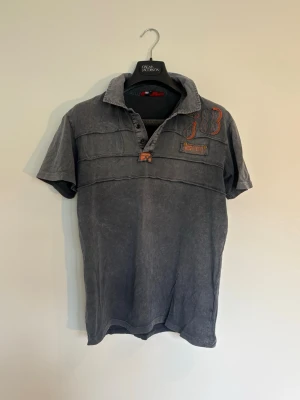 Grå pike från Diesel - Säljer en grå kortärmad piké från Diesel med orange broderade detaljer och tryck både fram och bak. Skjortan har knappar framtill och en avslappnad krage. Perfekt för dig som gillar en cool och avslappnad stil.