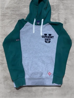 Superdry hoodie med gröna ärmar storlek m - Snygg hoodie från Superdry med grå kropp och gröna ärmar samt huva. Broderad Superdry Univ-logga på bröstet och klassisk känguruficka framtill. Vita dragsnören i huvan och ribbade muddar. Perfekt för en avslappnad streetstyle-look.