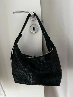 Svart flätad axelväska i läder - Snygg svart handväska med flätad design och justerbart axelband. Väskan har en silvrig dragkedja upptill och är tillverkad i läder. Perfekt för dig som gillar stilrena och trendiga accessoarer. 
