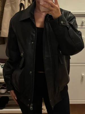 Svart oversized skinnjacka - Säljer en svart oversized skinnjacka med klassisk krage och dragkedja framtill. Jackan har två stora fickor med dragkedja och tryckknappar samt breda ärmslut. Perfekt för en cool och avslappnad stil.