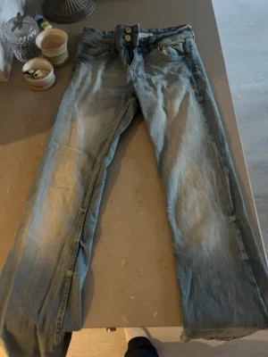 Ljusblå jeans med låg midja - Säljer ett par ljusblå jeans med hög midja och dubbla knappar framtill. Klassisk femficksmodell och raka ben. Jeansen har en snygg tvättad look och är perfekta för en avslappnad stil.