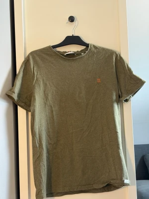Olivgrön t-shirt från Les Deux - Olivgrön t-shirt från Les Deux med liten broderad logga på bröstet. Klassisk rund halsringning och korta ärmar. Tillverkad i mjuk bomull för en skön känsla. Perfekt basic-plagg med stilren design.