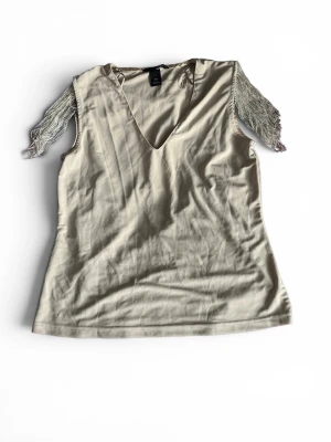 Beige topp med fransar och v-ringning - Snygg beige topp med v-ringning och glittriga fransar på axlarna. Tunn och mjuk i materialet, ärmlös och med en lös passform. Finns några pärlor som trillat av (se på bild 2)