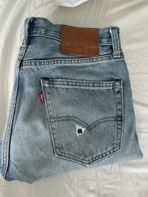 Levi's ljusblå raka jeans - Säljer ett par klassiska ljusblå jeans från Levi's med rak passform och femficksdesign. Jeansen har en snygg sliten look med ett litet hål på bakfickan och ikonisk brun läderpatch i midjan. Perfekta för en avslappnad stil. Storlek W 29 L 32