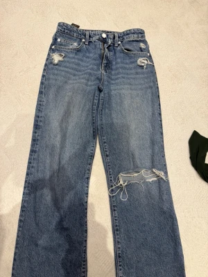 Blå slitna jeans från Mavi  - Säljer ett par blå jeans från Mavi  med raka ben och flera slitningar, bland annat ett stort hål på ena benet. Jeansen har klassisk femficksmodell och är tillverkade i denim. Perfekta för en avslappnad streetwear-look.