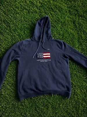 Mörkblå hoodie från Lexington - Snygg mörkblå hoodie från Lexington med broderad logga och amerikansk flagga på bröstet. Klassisk modell med huva och dragsko, samt mjukt material som känns skönt mot huden. Perfekt för en avslappnad och stilren look.