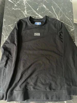 Svart sweatshirt från Jack & Jones - Säljer en svart sweatshirt från Jack & Jones med rund hals och diskret logga broderad på bröstet. Tröjan har ribbade muddar vid ärmslut och nederkant, och är gjord i mjukt bomullsmaterial. Perfekt för en avslappnad och stilren look.