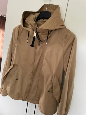 Beige vindjacka med huva Zara - Stilren beige vindjacka från Zara med huva och svarta dragskor med gulddetaljer. Jackan har dragkedja och tryckknappar framtill, samt två snedställda fickor med lock. Lätt material som passar perfekt för blåsiga dagar.