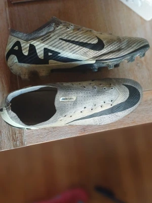 Nike Mercurial fotbollsskor  - Säljer ett par Nike Mercurial fotbollsskor i beige och svart med tydlig swoosh-logga på sidan. Skorna har låg profil, snörningslös design och mönstrad ovandel i syntetmaterial. Sulan har fasta dobbar och Zoom-teknologi för extra grepp på planen. Skorna är elite och är ganska använda så säljare dom billigt, storlek 43. Skorna är elite.