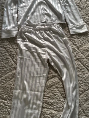 Vit satinpyjamas med ränder - Snygg vit pyjamas i satin med subtila vertikala ränder. Setet består av långärmad skjorta och byxor med resår i midjan. Perfekt för mysiga hemmakvällar och ser riktigt lyxig ut tack vare det glansiga materialet.