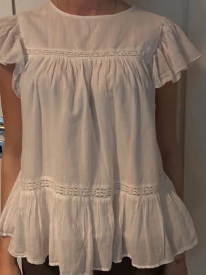 Vit volangblus med spetsdetaljer - Söt vit blus med volangärmar och spetsdetaljer. Blusen har en lös passform och är tillverkad i ett lätt och luftigt bomullstyg. Perfekt för en fräsch och somrig look.