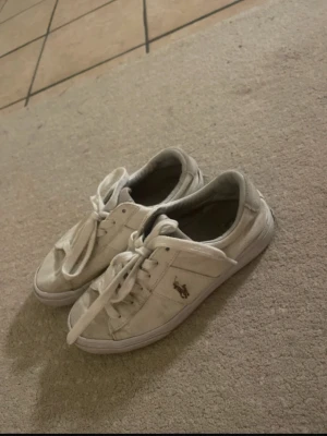 Vita låga sneakers från Polo Ralph Lauren - Klassiska vita sneakers från Polo Ralph Lauren med lågt skaft och snörning. Skorna har den ikoniska loggan broderad på sidan och är tillverkade i canvas med gummisula. Perfekta för en clean och avslappnad stil. Säljer på Vinted också mollyyii