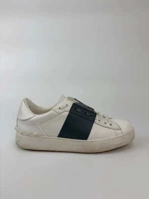 Valentino open skor - Snygga vita sneakers från Valentino Garavani med bred svart rem över mitten och nitar på hälen. Tillverkade i slätt läder med vita skosnören och låg profil. Perfekta för dig som gillar stilrena och exklusiva detaljer.