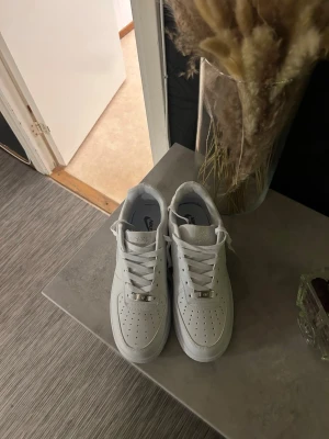 Vita Nike Air Force 1 sneakers - Säljer ett par klassiska vita Nike Air Force 1 sneakers i lågt utförande. Skorna har perforerad tå, vita skosnören och detaljer i metall framtill. Tillverkade i slätt läder med Nike-logga på sidan och plös. Perfekta för en clean och stilren look.