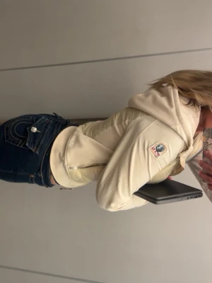 Ljusbeige dunväst från Parajumpers - Snygg ljusbeige dunväst från Parajumpers med quiltad design och dragkedja framtill. Västen har en huva och klassisk logotyp på ärmen. Perfekt för lager-på-lager och ger en sportig vibe till din outfit.
