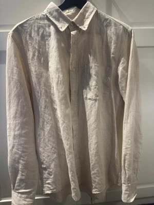 Beige linneskjorta med lång ärm - Stilren beige skjorta i linne med klassisk krage och bröstficka. Skjortan har knäppning framtill och långa ärmar, perfekt för en avslappnad men snygg look.