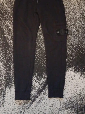 Svarta mjukisbyxor Stone Island - Säljer ett par svarta mjukisbyxor från Stone Island med ikonisk patch på benet. Byxorna har resår i midjan och vid bensluten samt en praktisk ficka på sidan. Perfekta för en avslappnad och trendig stil.