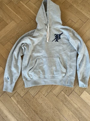 Grå hoodie från Polo Ralph Lauren - Snygg grå hoodie från Polo Ralph Lauren med stor huva och dragsko. På bröstet finns ett broderat P i mörkblå och på ärmen syns den klassiska lilla Polo-loggan. Hoodien har magficka och ribbade muddar. Perfekt för en chill och avslappnad stil.