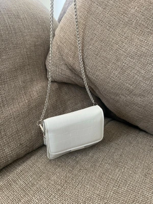 Vit axelväska med kedjerem - Snygg vit axelväska i croco-mönstrat konstläder med silverfärgad kedjerem. Väskan har en stilren, rektangulär form och stängs med lock. Perfekt för dig som gillar trendiga accessoarer med minimalistisk look.