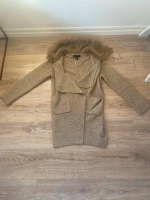 Beige cardiganjacka med fuskpäls - Mysig beige cardiganjacka från ONLY med stor krage i fuskpäls och två framfickor. Jackan har lång ärm och öppen front, perfekt för lager-på-lager-stil. Materialet är stickat och ger en avslappnad men ändå trendig look.