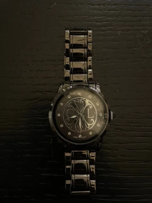 Svart klocka från Christian Lacroix - Snygg svart klocka från Christian Lacroix med metallarmband och dekorativa detaljer på urtavlan. Urtavlan har glittriga stenar som timmarkeringar och ett elegant mönster med CL-logga. Perfekt accessoar för att lyfta din stil.