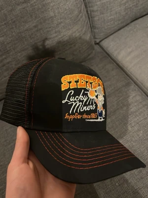 Svart Stetson keps Lucky Miners - Svart keps från Stetson med broderad text 'Lucky Miners' och en cool gubbe på framsidan. Kepsen har orange kontrastsömmar och mesh baktill för extra ventilation. Justerbar snapback för perfekt passform. Perfekt accessoar för en avslappnad streetstil.