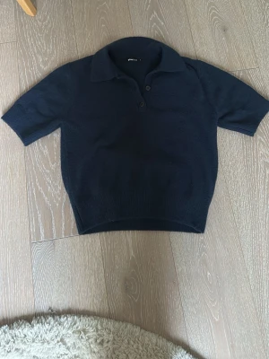 Mörkblå stickad pikétröja - Snygg mörkblå stickad pikétröja med korta ärmar och krage. Tröjan har två knappar framtill och ribbad nederkant. Perfekt för en stilren och avslappnad look.