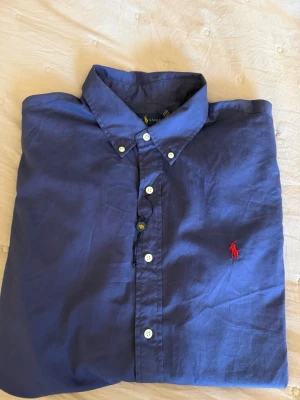 Aldrig använd Blå skjorta från Ralph Lauren - Klassisk blå skjorta från Ralph Lauren med vit knappstängning och röd broderad logga på bröstet. Skjortan är tillverkad i bomull och har en tidlös design med krage och långa ärmar. Perfekt för en stilren look. Aldrig använd, har varit hängt i garderob sen jag köpte den i 2022